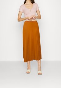 Haut court rose avec manches courtes à volants et trois boutons, associé à une jupe longue plissée couleur rouille et des sandales beige à enfiler.