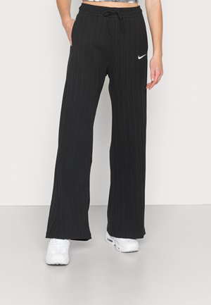 Person trägt schwarze, gerippte, weite Nike-Hose mit Kordelzug in der Taille und weiße Sneakers, steht vor einem schlichten Hintergrund.