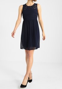 Marinblå ärmlös klänning med spetsliv, åtsittande midja och svängd kjol. Slät textur och knälång design. Bärs med svarta klackar.