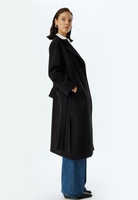 Een lange zwarte jas met oversized revers, zijzakken en een riem. Draag het over lichtblauwe wijde jeans met een gladde textuur.
