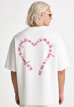 Personne aux cheveux clairs portant un t-shirt blanc avec un texte rouge en forme de cœur disant : Nous sommes des fleurs dans le même jardin, nous sommes des vagues dans la même mer.