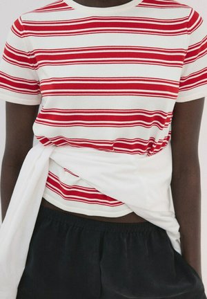 Persona dalla pelle scura che indossa una maglietta a maniche corte a righe rosse e bianche, pantaloni neri e un indumento bianco legato in vita.