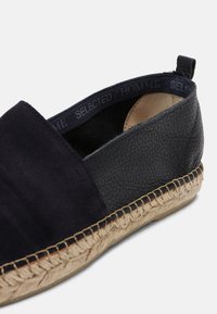 Chaussure mocassin en daim bleu marine et cuir texturé avec semelle espadrille en jute et détails cousus. L'intérieur présente une doublure en tissu foncé.