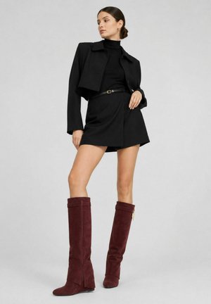 Mujer con chaqueta corta negra, cuello alto negro, falda negra con cinturón y botas altas de ante rojo oscuro, de pie contra un fondo gris.
