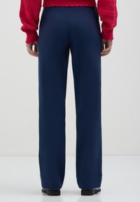 Personne portant un pantalon large bleu marine, des chaussures noires et un pull en laine rouge, se tenant devant un fond clair.