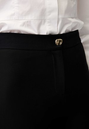 Schwarze High-Waist-Hose mit einem goldfarbenen Zierknopf, der oberhalb des Reißverschlusses befestigt ist, kombiniert mit einem in die Hose gesteckten weißen Hemd.