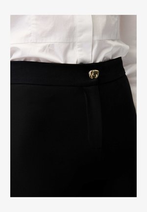 Zwarte hoge taille broek met een goudkleurige decoratieve knop boven de ritssluiting, gecombineerd met een ingestopt wit overhemd.