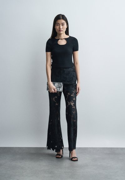 Γυναίκα που φοράει μαύρο τοπ με κοντά μανίκια και λεπτομέρεια με κλειδαρότρυπα, μαύρο παντελόνι από δαντέλα με floral σχέδιο, μαύρες γόβες, κρατώντας ένα μεταλλικό τσαντάκι σε απλό γκρι φόντο.