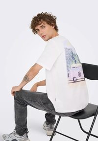 Joven con cabello rizado sentado en una silla negra, vistiendo una camiseta blanca con un estampado de furgoneta y bosque, jeans oscuros y zapatillas grises.