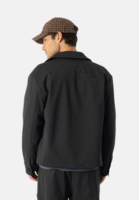 Ciemnoszara kurtka o dopasowanym kroju, z dużym kołnierzem i detalami z guzikami na rękawach, w połączeniu z brązową czapką w piedcę.