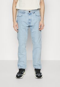 Jeans de mezclilla azul claro con un ajuste de pierna recta, cinco bolsillos y cierre de botón. Llevados con zapatillas deportivas negras y blancas.