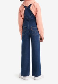 Tmavě modré džíny s nastavitelnými ramínky a dvěma zadními kapsami, v kombinaci s růžovou mikinou a bílými teniskami.