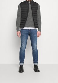 Svart quiltad väst över en grå strukturerad tröja och vit skjorta, i kombination med blå jeans och svarta ankelstövlar.