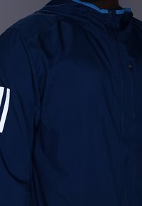 Chaqueta cortavientos azul marino con cremallera frontal, capucha y rayas blancas en la manga izquierda; material suave y ligero.