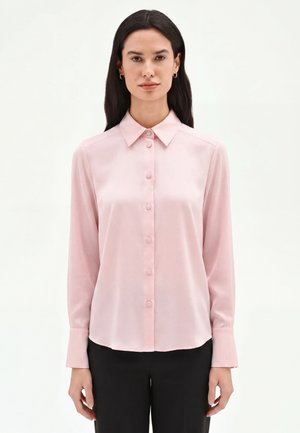 Femme aux longs cheveux foncés portant un chemisier rose clair boutonné et un pantalon noir, debout devant un fond blanc uni.