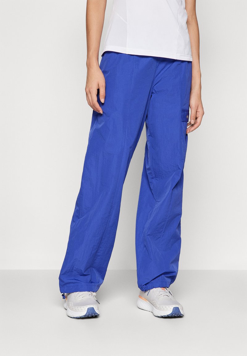 Calças soltas e azuis, confeccionadas em material leve, com um bolso tipo cargo na lateral e punhos elásticos nos tornozelos, usadas com sapatos cinzentos.