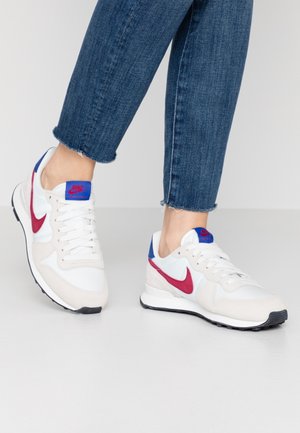 Person wearing white Nike sneakers with red swoosh and blue accents, paired with blue cropped jeans on a light gray floor.