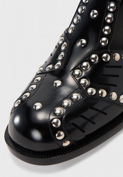 Botim de tornozelo em couro preto com tachas prateadas, bico arredondado, costura decorativa e sola de borracha com sulcos proeminentes.