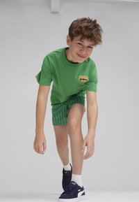 T-shirt verde in cotone con logo giallo, abbinato a pantaloni corti a righe verdi e sneakers blu navy. Design semplice con maniche corte e scollatura rotonda.