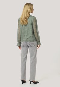 Blusa verde in pizzo con maniche lunghe, tessuto a motivo; abbinata a jeans grigi e tacchi neri, vista da dietro.