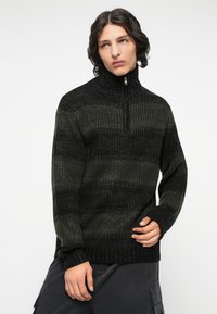 Dunkelgrüner und schwarzer strukturierter Strickpullover mit hohem Kragen und halbem Reißverschluss, der horizontale Streifenmuster und gerippte Bündchen aufweist.