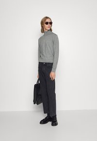Grå turtleneck genser, svarte avslappede jeans, svarte loafers, mørke solbriller og en svart veske, stående mot en enkel hvit bakgrunn.
