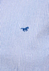 Light blue and white striped fabric with fine texture; features an embroidered dark blue dinosaur logo on the left side.