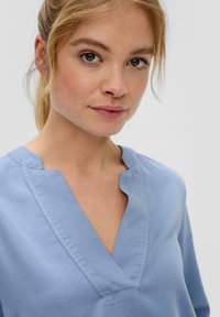 Helles Blau Bluse mit V-Ausschnitt und strukturiertem Kragen. Hergestellt aus glattem Stoff mit einer sauberen, minimalen Textur.