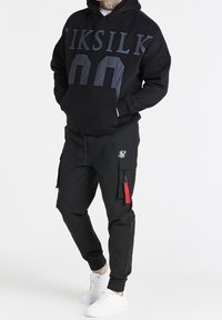 Felpa nera con la scritta "IKSKILK" e un grande numero. Abbinata a pantaloni cargo neri con tasche con zip e tirazip rossi. Sneakers bianche.