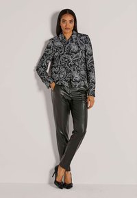Eine Frau steht an der Wand und trägt eine schwarz-weiße Paisley-Jacke, schwarze Lederhosen und schwarze hochhackige Schuhe mit Schleifen.