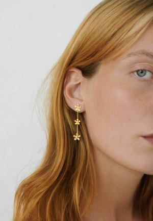 Donna con lunghi capelli rossi che indossa orecchini dorati pendenti con tre fiori collegati, su uno sfondo chiaro e semplice.