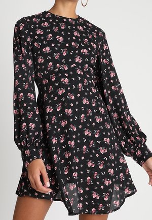 Kobieta w czarnej mini sukience z długim rękawem, ozdobionej różowym kwiatowym nadrukiem i rękawami z mankietami, stojąca na prostym jasnym tle.