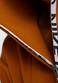 Veste orange zippée avec une fermeture éclair blanche ornée d'un motif de texte noir. Texture lisse avec un design à capuche et des détails de couture visibles.