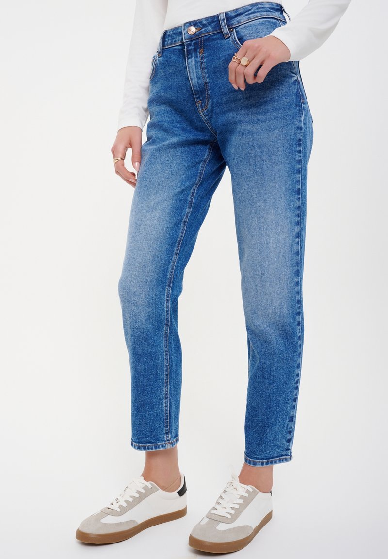 Kvinna klädd i blåblekta jeans, vit långärmad topp, vit och grå sneakers, och ringar på fingrarna mot en vit bakgrund.