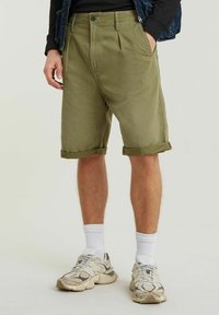 Pantaloni corti in cotone verde oliva con una vestibilità rilassata, caratterizzati da risvolti e pieghe frontali. Abbinati a calzini bianchi e sneakers chiare.
