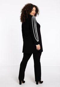 Tunique sportive noire avec un design à trois rayures blanches sur les manches, associée à un pantalon noir et des talons, présentant une coupe décontractée et une texture lisse.