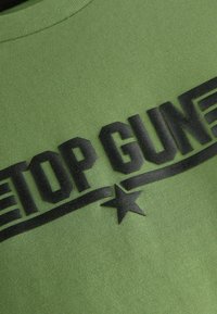 Zelené bavlněné tričko s černým potiskem "TOP GUN" obsahující hvězdu a vodorovné pruhy, žebrovaný kulatý výstřih a hladkou texturu.