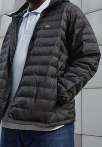 Man med svart quiltad jacka från Levi's över en ljusgrå polo och blå jeans, stående mot en ljusgrå kaklad vägg.