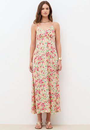 Femme debout portant une robe sans manches à motif floral, longue jusqu'aux chevilles avec de fines bretelles, associée à des sandales dorées et des bijoux.