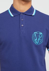 Polo navy avec une bordure turquoise contrastante sur le col et les manches. Présente un logo brodé turquoise décoratif sur la poitrine.