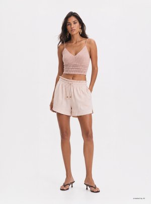 Donna che indossa un top corto rosa all’uncinetto, pantaloncini beige chiaro con coulisse e sandali neri con tacco, in piedi davanti a uno sfondo bianco semplice.
