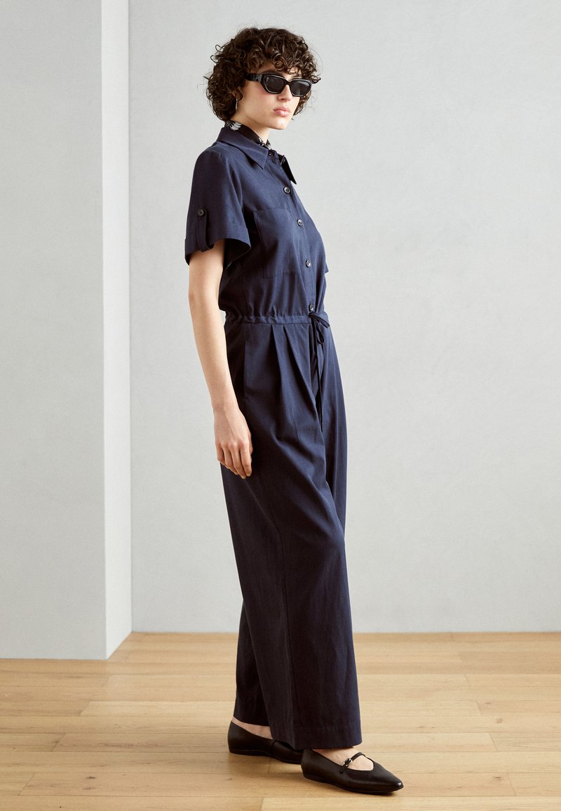 Frau mit kurzen lockigen Haaren, die schwarze Sonnenbrillen, einen navyblauen Kurzarm-Jumpsuit mit Knöpfen und schwarze flache Schuhe trägt, steht drinnen.