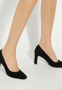 Legs wearing black suede mid-heel pumps on a white background, showing the shoes from side and front angles.