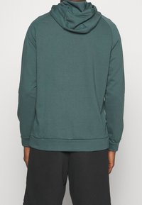Sweat à capuche en teal, fabriqué dans un matériau doux ; il dispose d'une poche kangourou, de poignets côtelés et d'une coupe décontractée. Texture lisse, sans motifs visibles.