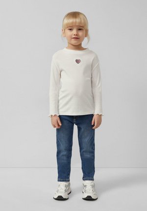 Jeune fille aux cheveux blonds portant un chemisier blanc à manches longues avec un petit motif en forme de cœur, un jean bleu et des baskets blanches, debout.