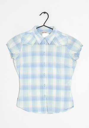 Vaaleansininen ja valkoinen ruudullinen lyhythihainen nappipaita, jossa on terävä kaulus ja röyhelöidyt hihansuut, ripustettuna mustaan henkarille valkoista taustaa vasten.