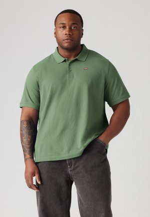 Groene poloshirt met korte mouwen, gemaakt van een gladde stof. Heeft een klein rood logo op de borst en een klassieke kraag. Draagt met een paar grijze jeans.