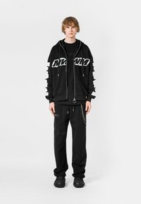 Sudadera negra con cremallera y gráficos blancos, combinada con una camiseta negra. Acompañada de pantalones negros con detalles de cadena y zapatos de goma.