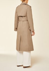Femme vue de dos portant un long trench-coat beige, un pantalon blanc à jambes larges et des chaussures marron à talons sur un fond uni.