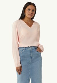 Rosa Langarmbluse mit V-Ausschnitt und gerafften Ärmelbündchen, kombiniert mit einem hellblauen Jeansrock mit seitlichem Schlitz.